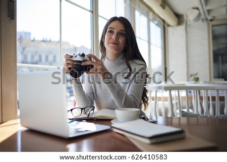Young attractive female journalist testing work of vintage camera preparing for retro photo session sitting in coffee shop during work break,skilled photographer taking picture of modern cafe interior
