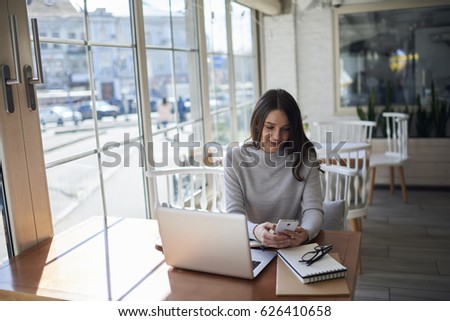 Attractive female editor reading notification on modern smartphone while working online updating multimedia files on website, charming owner of business using application on cellular to share files
