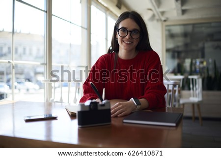 Portrait of young teen female student of college dressed in casual outfit and eyewear  making homework task sitting in coworking space, talented writer creating article in good mood during leisure
