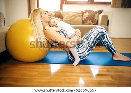 Blonde mother holding her sleeping toddler daughter in arms while relaxing on fitness ball after home workout exercise. Toned image. Authentic people.