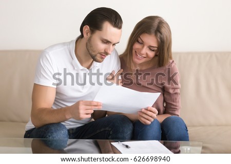 Smiling couple holding papers and carefully studying documents sitting on sofa indoors, reading terms and conditions, reviewing agreement, considering mortgage loan offer, personal insurance  Royalty-Free Stock Photo #626698940