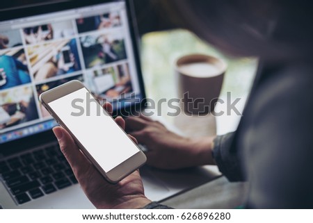 Mockup image of a business woman holding mobile phone with blank white screen while using laptop with coffee cup on the table in office