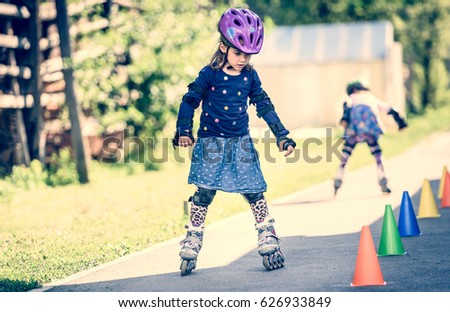 Children learning to roller skate on the road with cones. Twin girls are practising safe roller skating on a home driveway road wearing protective gear - helmets, knee, elbow and hand protectors pads.