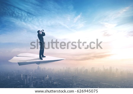 Picture of a young businessman standing on a paper plane while flying above a city and looking through a binoculars