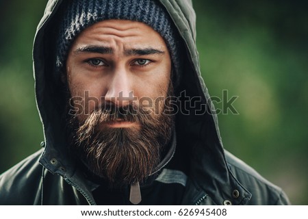 Outdor porttrait of a handsome brutal bearded man with dark beard and mustache dressed in winter clothes, covered with frost. Man wear coat hat and hood. look like hipster or tourist. Close up.