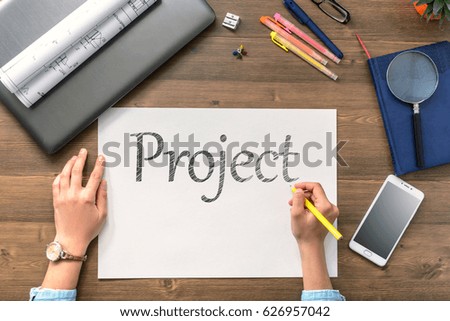 The girl sits at the table with a mobile phone, a laptop, business accessories and a sheet with text Project