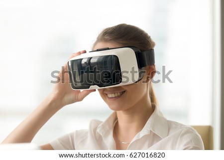 Smiling young woman looking in virtual reality glasses. Female enjoying VR technology digital simulation. Girl entertains with electronics 3d goggles. Virtually conversation with colleagues or friends