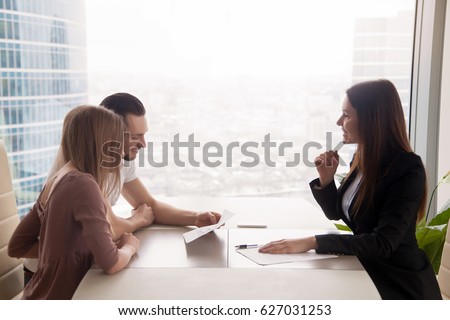 Side view of realtor and young couple sitting at office desk discussing property for sale. Potential buyers holding house plan, considering investment in home or taking loan to purchase real estate  Royalty-Free Stock Photo #627031253