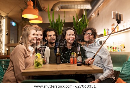 leisure, technology, friendship, people and holidays concept - happy friends with drinks and taking picture by smartphone selfie stick at bar or cafe
