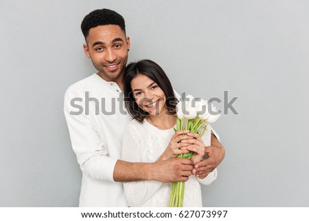 Image of young happy loving couple standing over grey wall with flowers. Looking at camera.