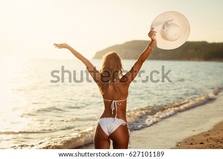 Beautiful young woman in swimsuit holding summer hat enjoying at sunset on the beach. Rear view. 