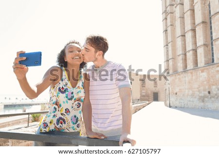 Young ethnically diverse couple sightseeing in destination city cathedral monument, kissing, holding a smart phone taking selfies photos on holiday, outdoors. Travel technology lifestyle recreation.