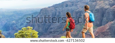 Hikers couple hiking in mountains landscape, banner panorama. Woman and man walking on hike in Waimea Canyon State Park, Kauai, Hawaii, USA. Looking at view happy enjoying healthy outdoor lifestyle.
