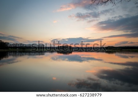 Horizontal photo with spring evening landscape after sunset. Cloudy sky is nicely reflected in the water of small pond. Grass, reed, threes and branches are in background and foreground.