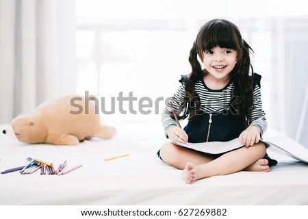 Adorable little Asian girl sitting on her bed playing drawing coloring book while looking aside smiling with happiness in natural sunlight tone with copy space to the left, concept children activity.