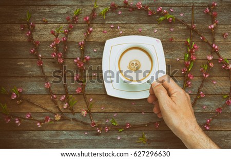 Cup with coffee milk in a male hand on a background of an old wooden table with peach branches with pink flowers, top view. Retro toning photo with vignette