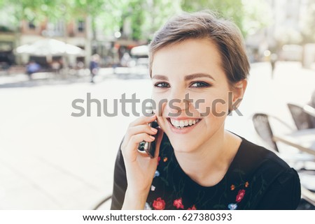 Close-up portrait of attractive young Caucasian woman talking on mobile phone at outdoor cafe. Girl talking by mobile phone, smiling. Sunny day. Outdoor photo.