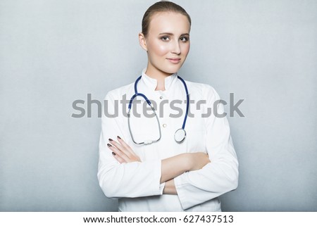 Portrait of young female confident caucasian doctor physician with makeup in white gown with stethoscope on her neck and crossed arms over her chest on white background in studio or medical center
