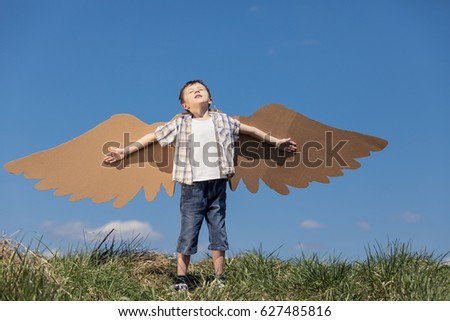 Little boy playing with cardboard toy wings in the park at the day time. Concept of happy game. Child having fun outdoors. Picture made on the background of blue sky.