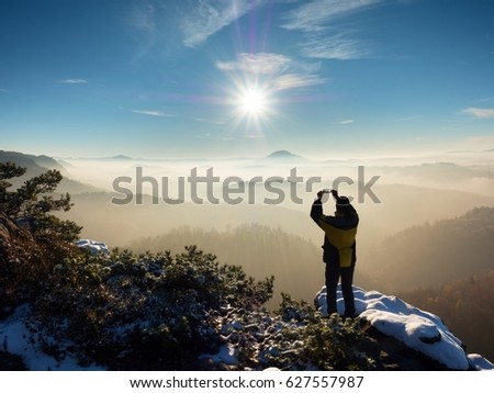 Hiker hold phone above head, take picture of misty winter landscape. Exposed rocks covered with fresh powder snow. Stony rock peak increased from foggy valley. 