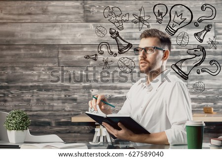 Attractive young man writing in notepad at workplace with coffee cup, supplies, other items and creative sketch. Strategy concept