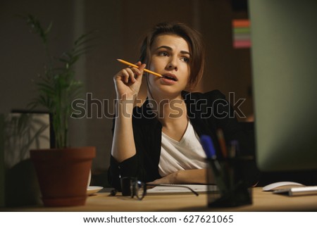 Picture of serious young lady designer sitting in office at night using computer. Looking aside holding pencil.