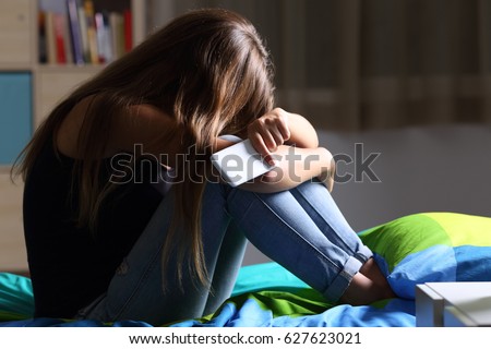 Single sad teen holding a mobile phone lamenting sitting on the bed in her bedroom with a dark light in the background Royalty-Free Stock Photo #627623021