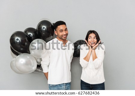 Image of happy young loving couple standing over grey wall and looking at camera. Man holding a lot of balloons.