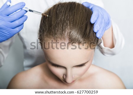 Doctor aesthetician in blue medical gloves and white medical gown makes hyaluronic acid rejuvenation beauty injections in the head of female patient for hair growth and to prevent boldness. Upper view