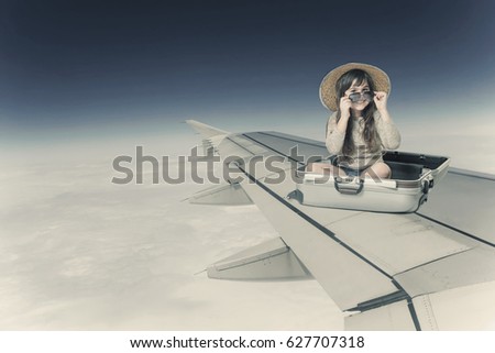 Smiling little girl in glasses and hat is sitting in the open suitcase on the wing of the plane in flight. Edited as a vintage photo.