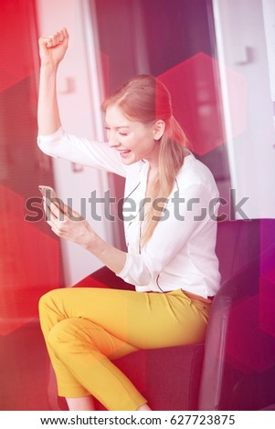 Smiling young businesswoman cheering while holding mobile phone on chair in office