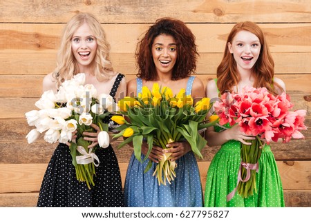 Three surprised women in dresses posing near the wooden wall and holding bouquets of flowers  while looking at the camera