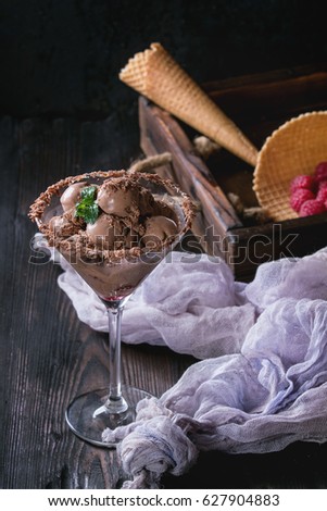 Cocktail glass with chocolate ice cream mini balls with fresh raspberries, mint and waffle cone in wood tray with textile gauze over black burnt wooden background.