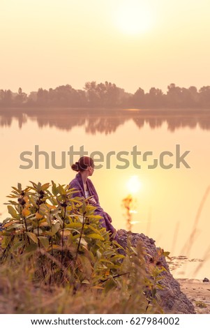 The girl meets the dawn on the lake. She is turned with her back. The woman wrapped herself in a blanket.