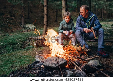 Father with son warm near campfire, drink tea and have conversation Royalty-Free Stock Photo #628027658