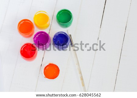 Open jars with colorful paint and transparent clean brush on a white wooden background