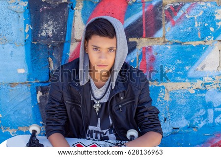 Stylish teenager boy wearing hood shirt with skateboard in city graffiti background looking at you camera sitting