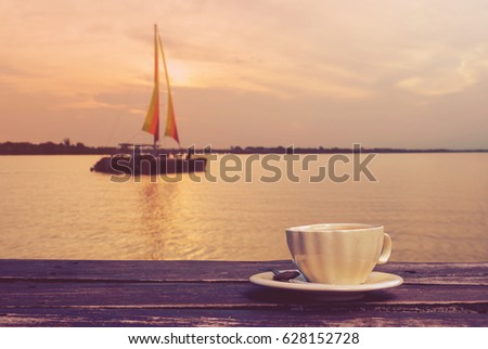a cup of hot coffee on wooden table with a blurry sail boat in a lake and the evening sky background, filtered tones