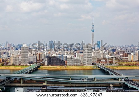 A panoramic view of Tokyo City skyline under sunny sky, with bridges over Arakawa River, elevated expressways along the riverside & famous landmark Tokyo Skytree among crowded buildings in background