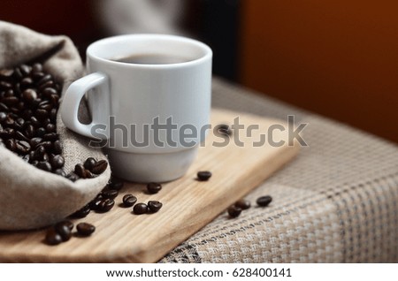 A still life with a bag full of brown coffee beans, which lies next to a small white cup of hot coffee on a wooden board behind a small low table covered with a checkered tablecloth