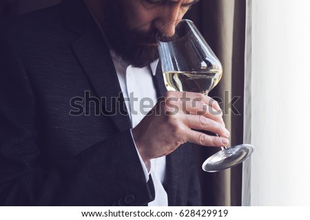 horizontal close up of a Caucasian man with beard black suit and white shirt tasting a glass of white wine by the window natural lighting Royalty-Free Stock Photo #628429919