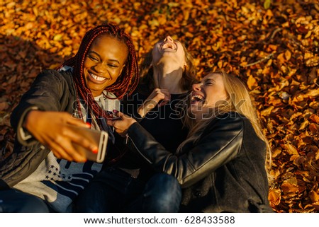 very happy female friends making funny selfie in autumn park. Cute girls with different colored skin. Female lying in yellow leaves and making smiling faces at camera. Sunny autamn day
