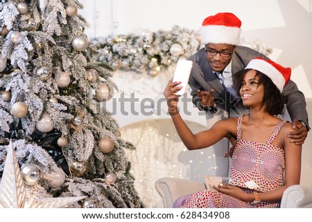 The beautiful afro-american couple in christmas caps smiling and making selfies near the Christmas Tree