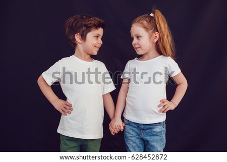 Two children in white t-shirts stand holding hands on black background. Mock up. Royalty-Free Stock Photo #628452872