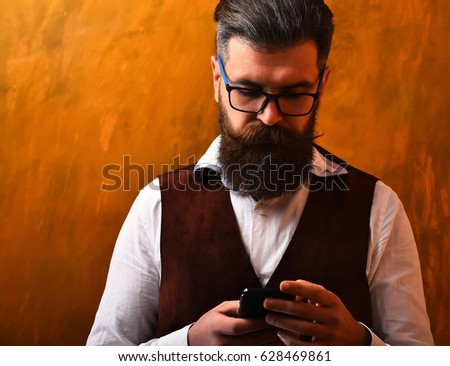 Bearded man, long beard. Brutal caucasian unshaven serious hipster with glasses and moustache surfing internet on cell phone, wearing white shirt and suede waistcoat on brown studio background