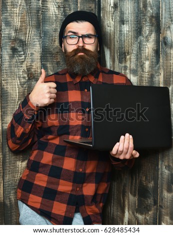 Bearded man, long beard. Brutal caucasian serious unshaven hipster holding laptop in red black checkered shirt with hat and glasses on brown vintage wooden studio background