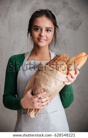 Picture of young happy lady baker standing and holding bread. Looking at camera.