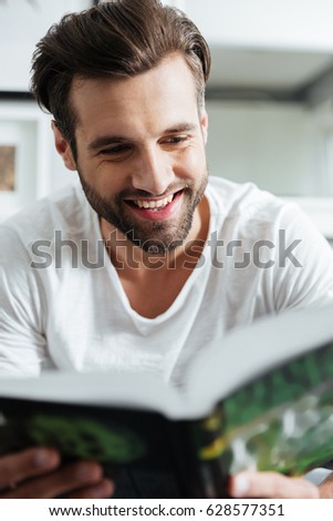 Picture of young smiling man sitting on sofa indoors at home while reading book. Looking aside.
