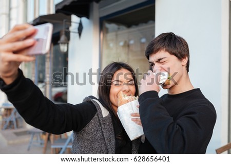 Brother and sister making selfie with closed eyes while eating burgers in the street