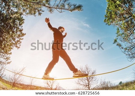 A man, aged with a beard and wearing sunglasses, balances on a slackline in the open air between two trees at sunset Royalty-Free Stock Photo #628725656